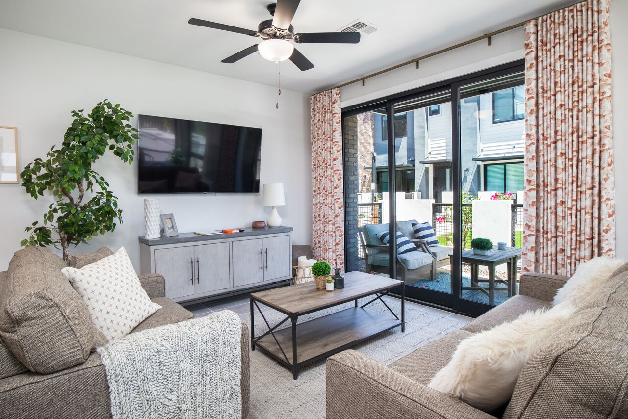 A modern living room with a ceiling fan, a flat-screen TV, a grey sofa, a coffee table, and a sliding glass door leading to an outdoor patio.
