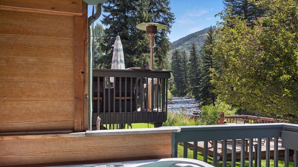 A wooden deck with a hot tub, a covered seating area, and a view of a river and forested mountains. - Deck with Private Hot Tub - Updated Townhome Overlooking Gore Creek + Parking for 4 (Vail)
