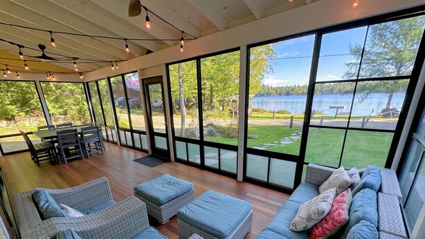 Una habitación amplia con muebles de mimbre, ventanas grandes que dan a un lago y follaje, y un ventilador de techo.