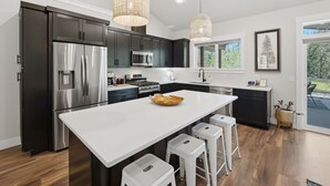 A modern kitchen with dark cabinets, stainless steel appliances, a large island with stools, and pendant lighting. - Clark Fork Crossing - Adventures You Seek in Western Montana.  River access.  (Superior)