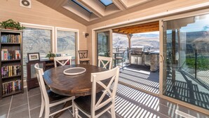 A dining area with a wooden table and chairs, a bookshelf, and a view of a mountainous landscape through large glass doors. - Riverview Pool Villa near Chelan, and Hot Tub! (Chelan)