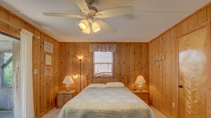 A bedroom with a wooden paneled wall, a bed with a white bedspread, two bedside lamps, a ceiling fan, and a window with blinds. - Oceanfront, Amazing Sunrises, Dog Friendly, Private deck & Screened in Porch (Avon)
