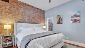 A bedroom with a brick wall, a white bed, two bedside tables with lamps, and two framed pictures on the wall. - Modern Broughton Loft with Southern Charm (Savannah)