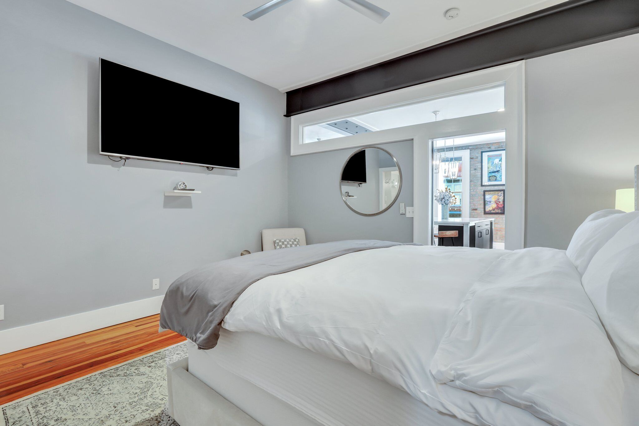 A modern bedroom with a large bed, a flat-screen TV mounted on the wall, a round mirror, and a small desk with a chair.