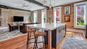 A modern kitchen with a large island, bar stools, and a dining area with a brick wall and large windows. - Modern Broughton Loft with Southern Charm (Savannah)
