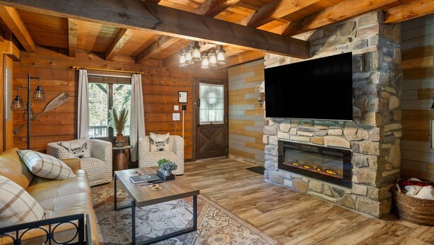 A cozy log cabin interior with a stone fireplace, wooden ceiling beams, and a large flat-screen TV mounted on the wall. - Rustic Luxury - Log Cabin in the Pocono Mountains - Indoor Sauna- Close to Ski (Pocono Lake)