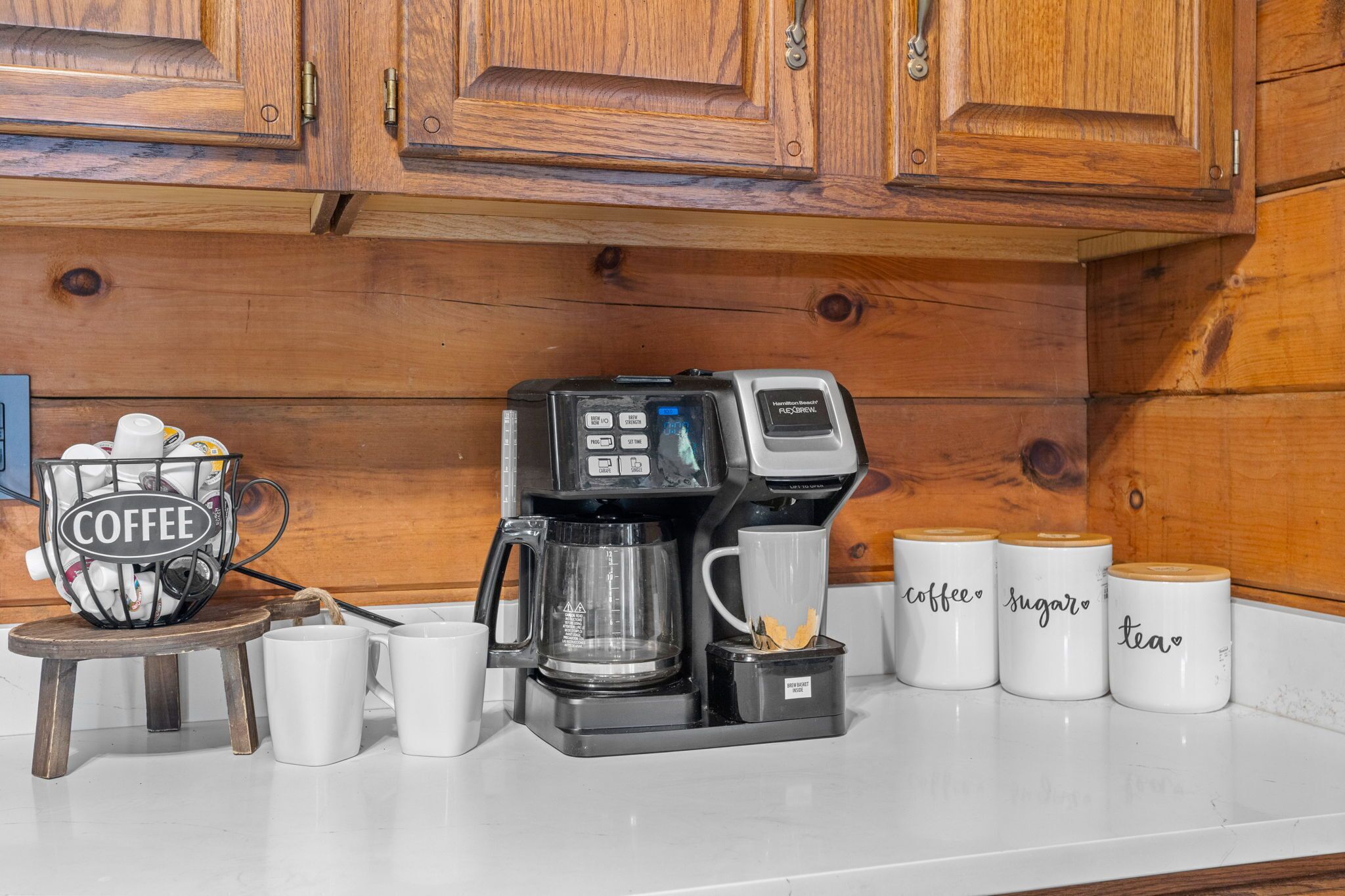 A coffee maker on a kitchen counter with a coffee basket and labeled mugs for coffee, sugar, and tea.