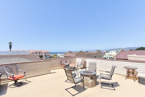 A rooftop terrace with outdoor furniture, a stone fire pit, and a view of the ocean and buildings. - Large home Steps from the Sand (Oceano)