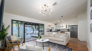 Una sala de estar moderna con un sofá, sillones y una mesa de centro. Se ve una puerta corrediza de vidrio que da a un balcón con mobiliario exterior. En el fondo, se observa una cocina con gabinetes blancos y un refrigerador.