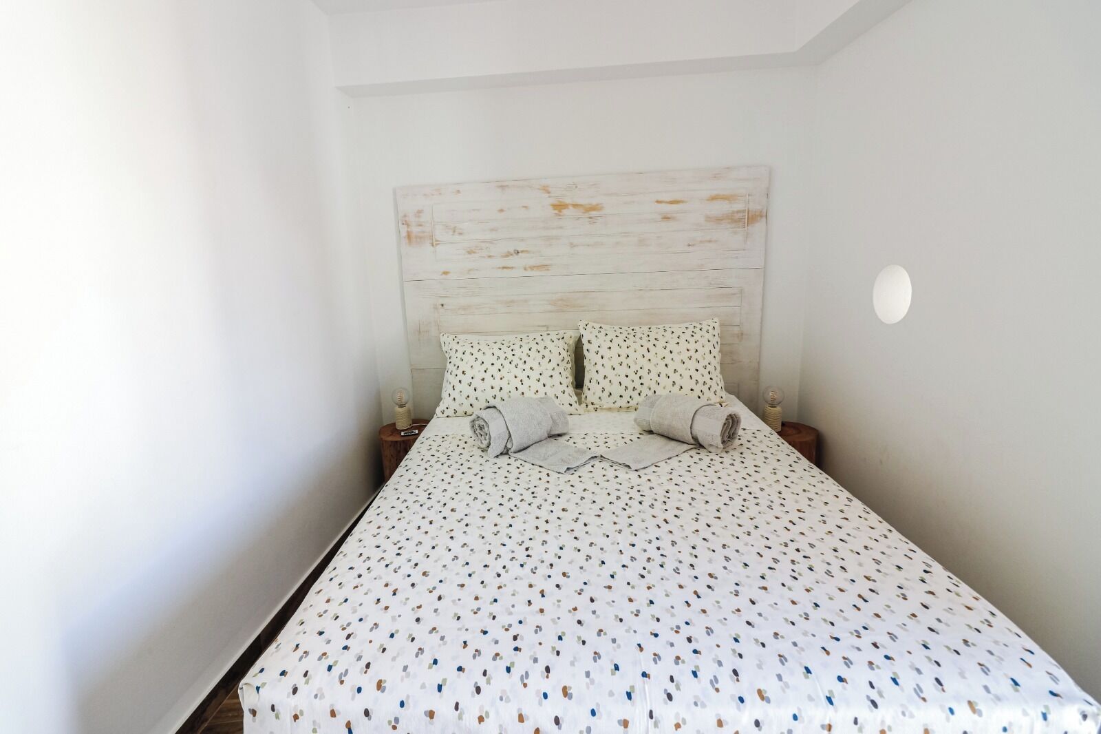 A neatly made bed with a patterned bedspread and pillows, a wooden headboard, and a small round nightstand with a lamp.