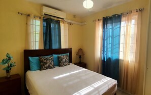 A hotel room with a bed, an air conditioning unit, a nightstand, a lamp, and curtains. - Mandarin Vacation Villa 2 Bedrooms - Toco (Toco)