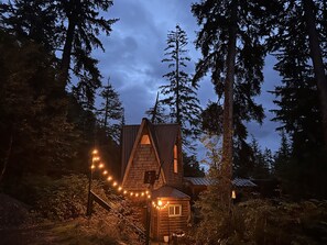 A cabin surrounded by tall trees, with string lights leading to the entrance. - Organic-mod mid-century A-frame gem in pristine coastal forest setting (Juneau)