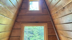 A wooden interior with a sloped ceiling, a hanging light fixture, and two windows providing views of greenery.