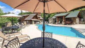 A poolside area with lounge chairs and umbrellas, surrounded by wooden cabins with stone walls.