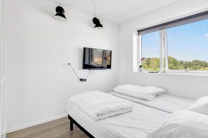 A bedroom with a TV mounted on the wall, a window with a view of greenery, and a bed with white bedding.