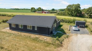 A black building with a tiled roof, a car, and a small shed with trash bins. - New and modern holiday home with hot tub (Frørup)