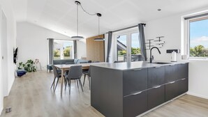 A modern kitchen with a large island, dining area with chairs, and a view through large windows. - New and modern holiday home with hot tub (Frørup)