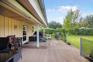 A covered outdoor deck with seating, a grill, and a view of a well-maintained lawn and trees.