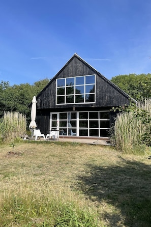 A modern building with a large window, a door, and a small outdoor seating area with a white chair and umbrella. - Lovely beach home enveloped in green surroundings (Ebeltoft)
