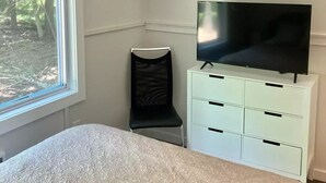 A bedroom with a bed, a TV on a white dresser, a window with blinds, and a ceiling light fixture.