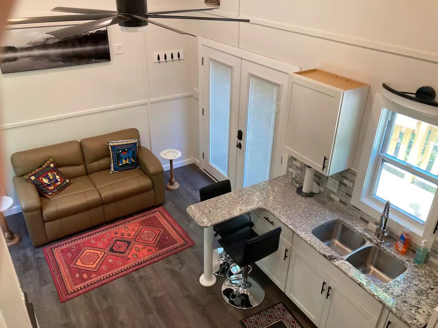 Un espacio habitable compacto con kitchenette, un sofá de cuero marrón con cojines estampados, una alfombra roja y un ventilador de techo.