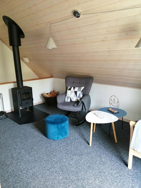 Une pièce chaleureuse avec un poêle en bois noir, un pouf bleu, un fauteuil gris agrémenté d’un coussin à motifs et une petite table blanche ornée d’objets décoratifs.