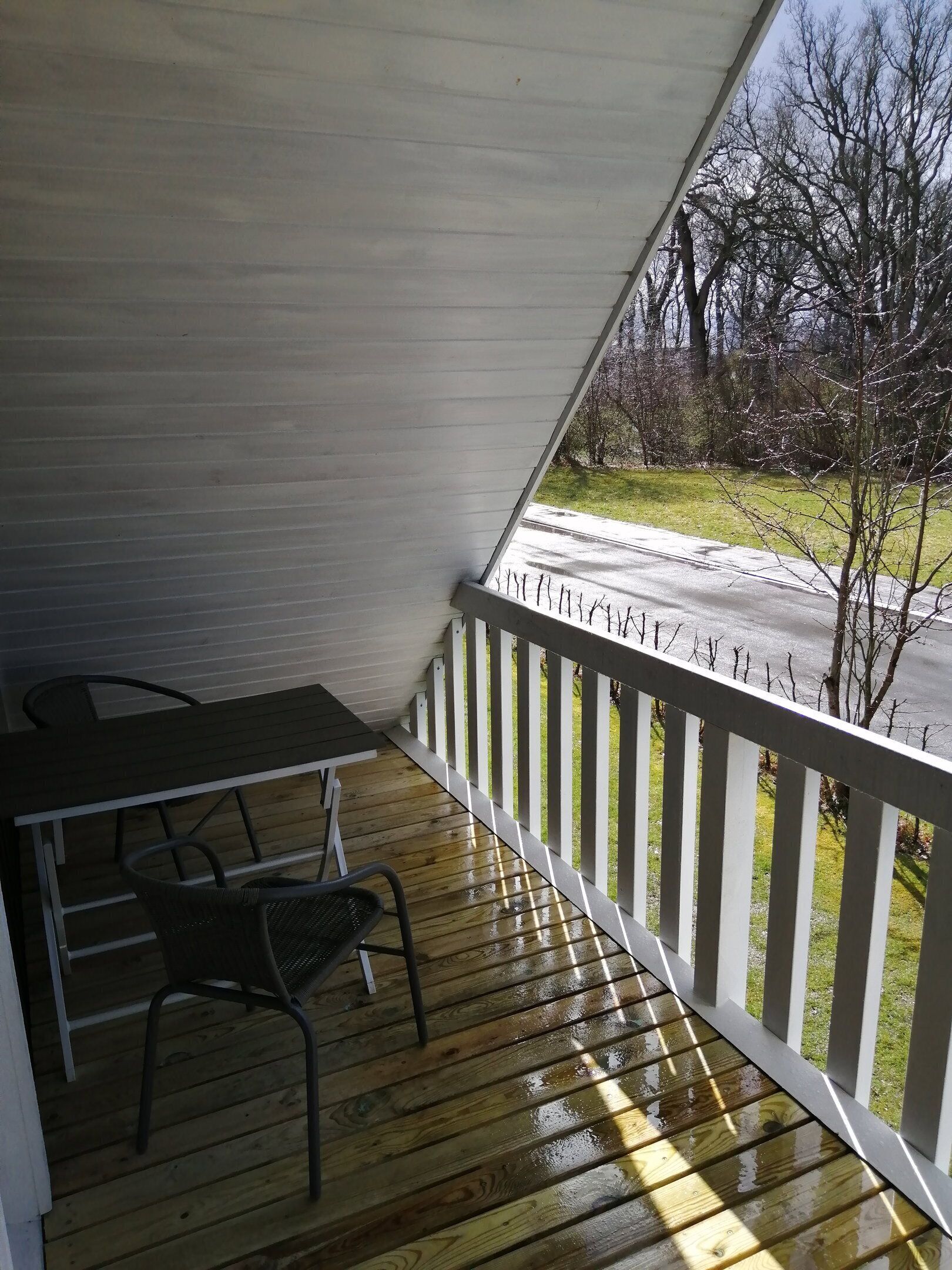 A covered porch with a table and chairs, a white railing, and a view of a road and trees.
