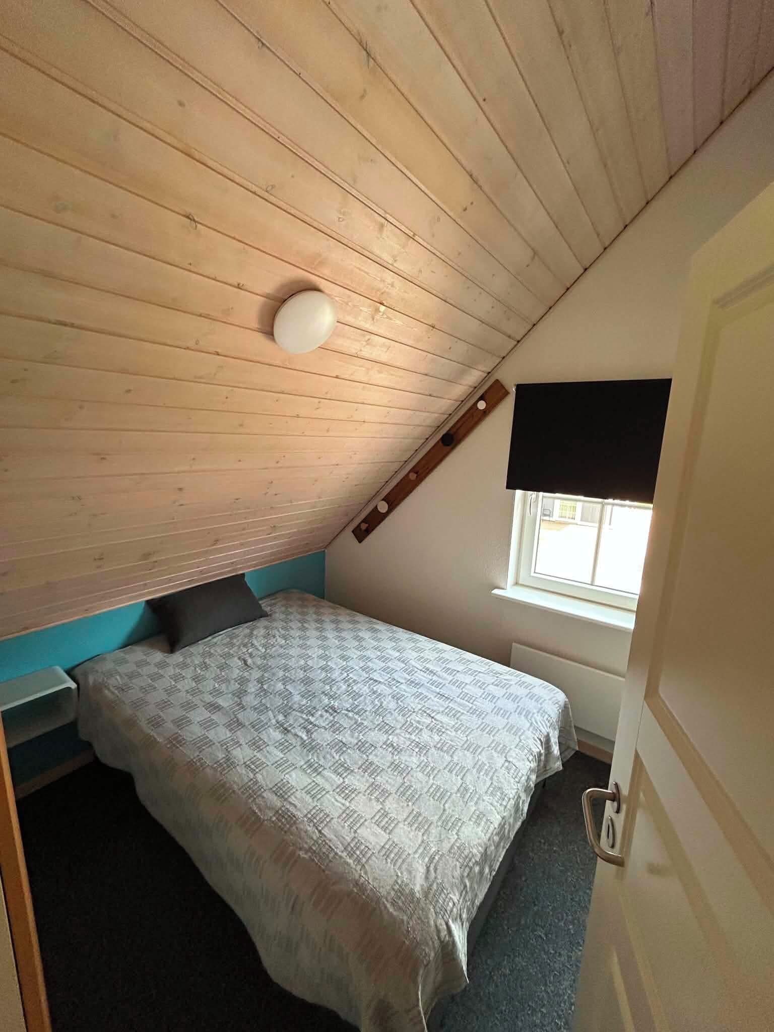 A small, single-bed room with a sloped ceiling and a window with a blind.