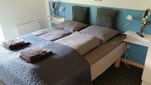 A neatly made bed with a blue quilt, a grey blanket, and two brown towels. There are wall art pieces depicting cities, a white bedside table with a lamp, and a small vase with flowers. - Summer house for relaxation and activity (Gråsten)