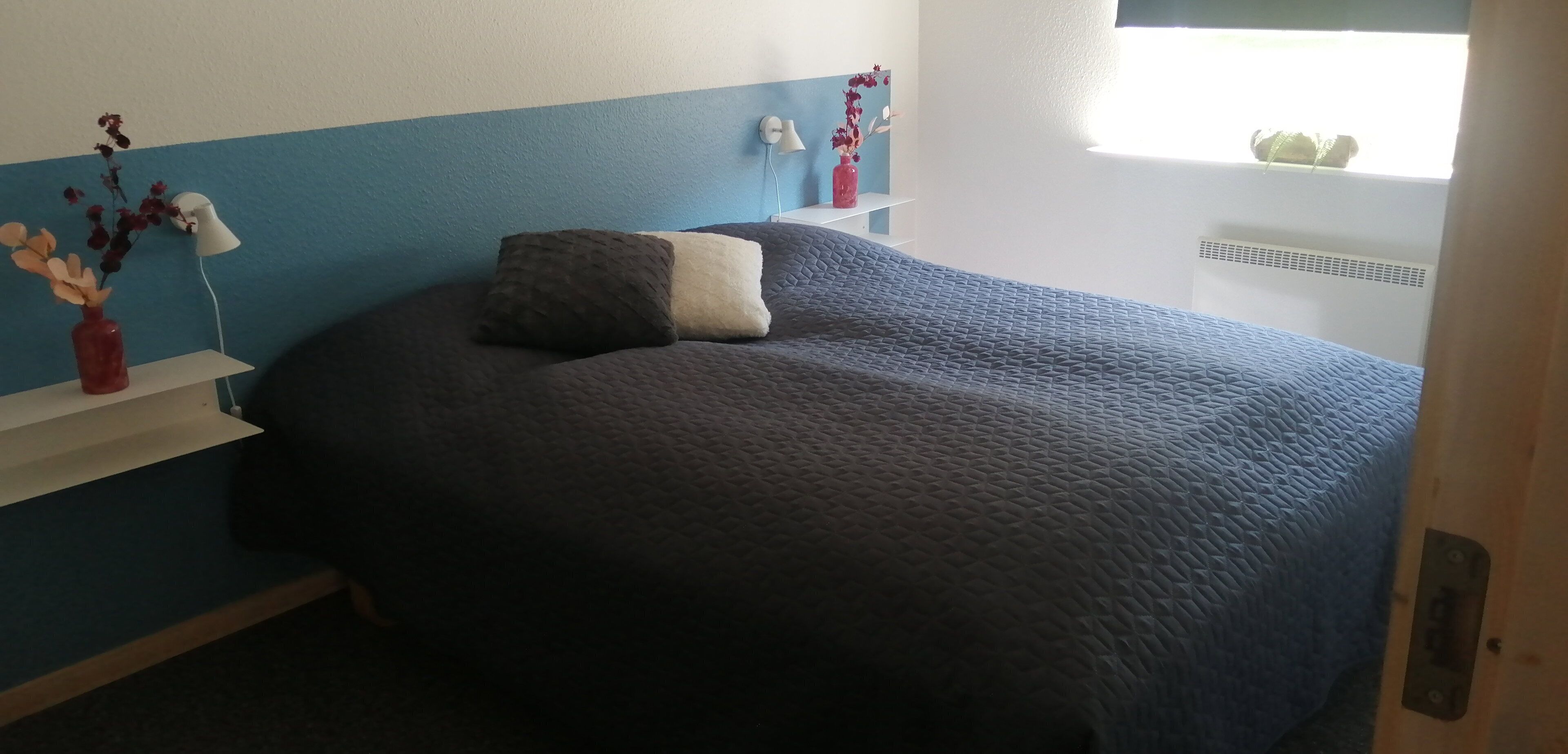 A bedroom with a bed, a nightstand, a lamp, a vase with flowers, and a window.