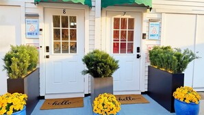 Deux portes, portant les numéros 8 et 7, chacune agrémentée d’une pancarte « BIENVENUE », de plantes en pot et d’un paillasson avec l’inscription « bonjour ».