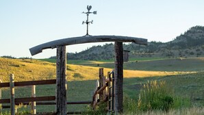 A rustic wooden structure with a weather vane, surrounded by a wooden fence and a grassy field with a hill in the background.