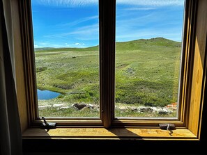 Une fenêtre donnant sur un paysage verdoyant, un petit plan d’eau et des collines lointaines.