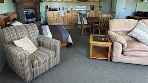 A cozy living room with a striped armchair, a sofa, and a glass coffee table. A kitchen with wooden cabinets and a refrigerator is visible in the background.