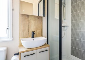 Un baño con lavamanos blanco, tocador de madera, cabina de ducha con mampara de vidrio y un inodoro blanco.