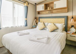 Una cama bien hecha con almohadas, una mesita de noche con un reloj, una estantería con adornos y una ventana con cortinas.