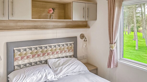 A bedroom with a bed, a nightstand, a wall-mounted lamp, a window with curtains, and a wooden shelf with a vase.
