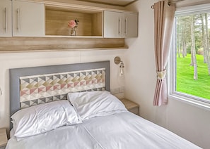 A bedroom with a bed, a nightstand, a wall-mounted lamp, a window with curtains, and a wooden shelf with a vase. - This stunning holiday home with a double bedroom and en suite W. (Bishop Auckland)