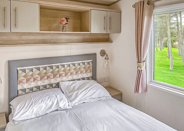 Un dormitorio con cama, mesita de noche, lámpara de pared, ventana con cortinas y estante de madera con un jarrón.
