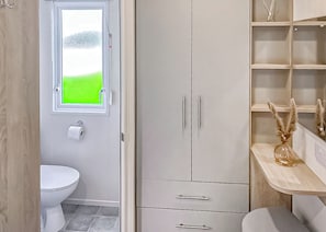A bathroom with a toilet, a window with a green view, a white wardrobe, and a shelf with decorative items. - This stunning holiday home with a double bedroom and en suite W. (Bishop Auckland)