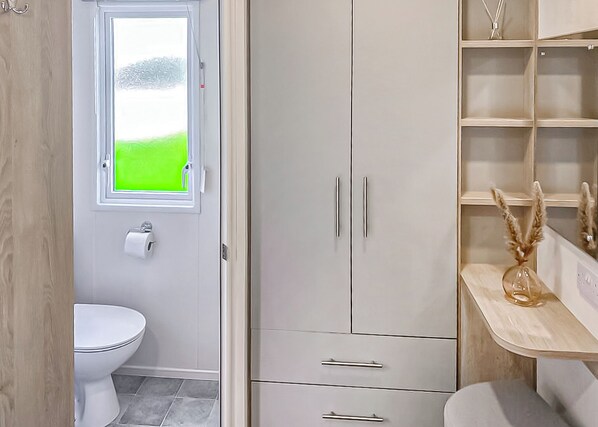A bathroom with a toilet, a window with a green view, a white wardrobe, and a shelf with decorative items. - This stunning holiday home with a double bedroom and en suite W. (Bishop Auckland)