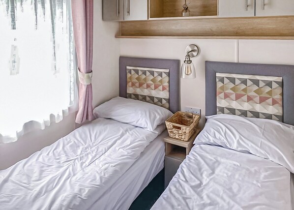 A bedroom with two single beds, a wooden headboard, a bedside table with a basket, and a window with curtains.