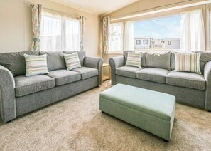 A living room with two grey sofas, matching cushions, a green ottoman, and a view of a residential area through large windows.