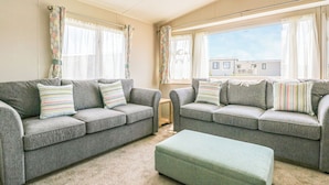 A living room with two grey sofas, matching cushions, a green ottoman, and a view of a residential area through large windows.