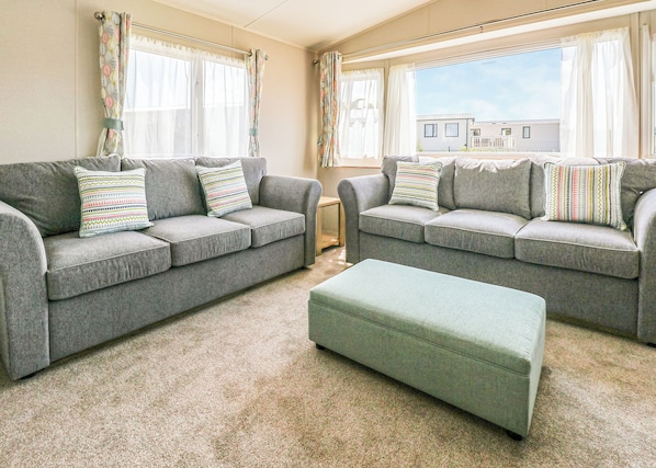 A living room with two grey sofas, matching cushions, a green ottoman, and a view of a residential area through large windows.