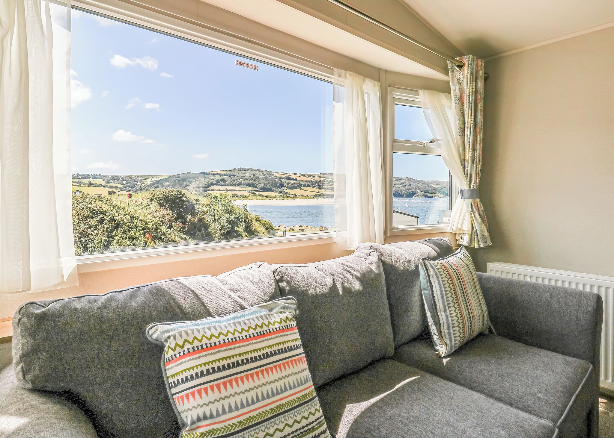 Quarto com sofá cinzento, almofadas estampadas e vista para a paisagem costeira através de grandes janelas.