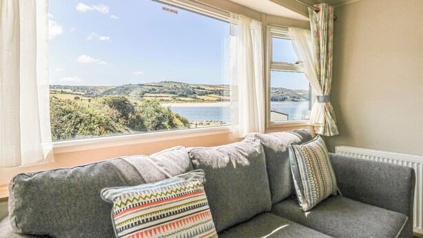 A room with a grey sofa, patterned pillows, and a view of a coastal landscape through large windows.