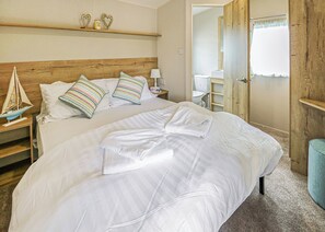 A neatly made bed with white linens, a wooden headboard, and a small shelf with decorative items. - Large 12ft-wide caravan with one double with en suite W. (Cardigan)