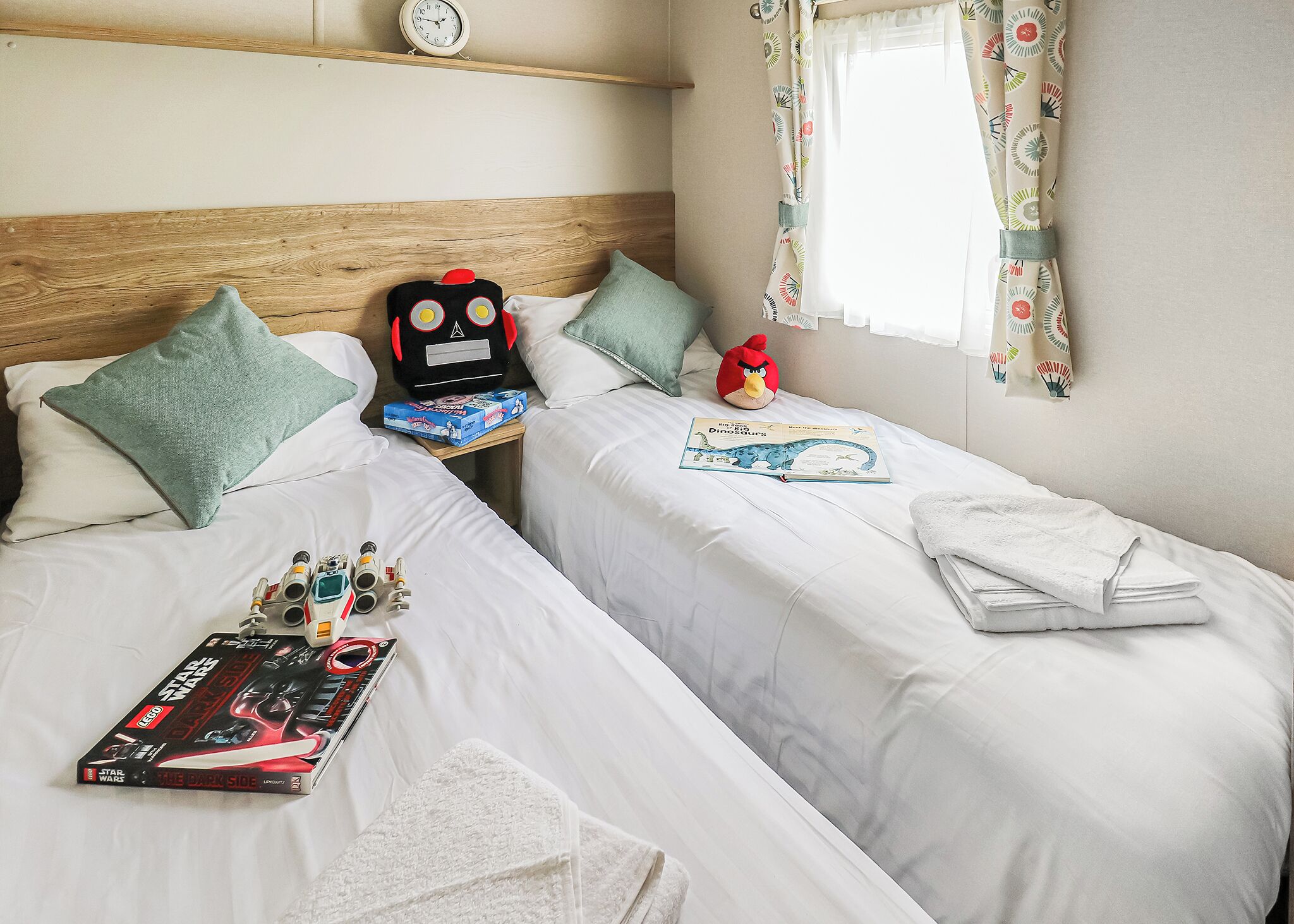 Une chambre d’hôtel avec deux lits, une horloge au mur, une fenêtre avec des rideaux et des jouets sur les lits.