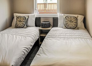 A bedroom with two beds, each with a clock pillow, a small table between them, and a window with a view of a balcony.
